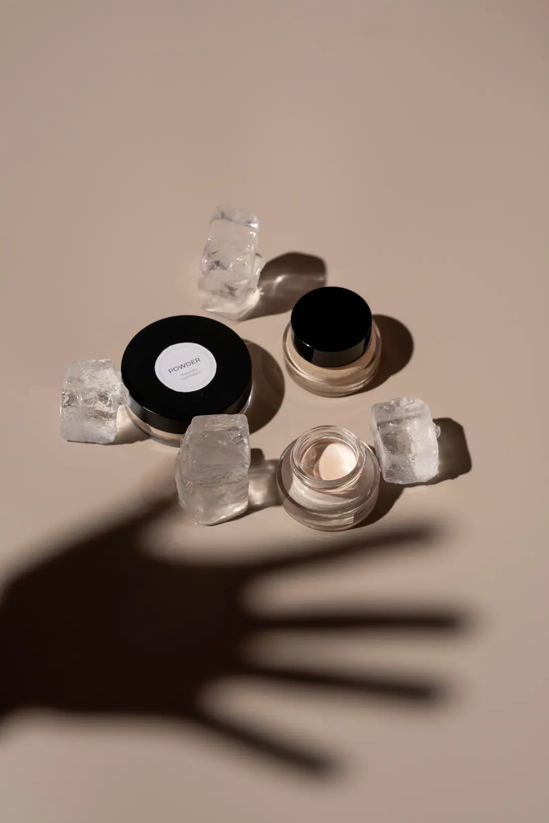 Cosmetic jars and clear crystals on a taupe surface beneath the shadow of an open hand.